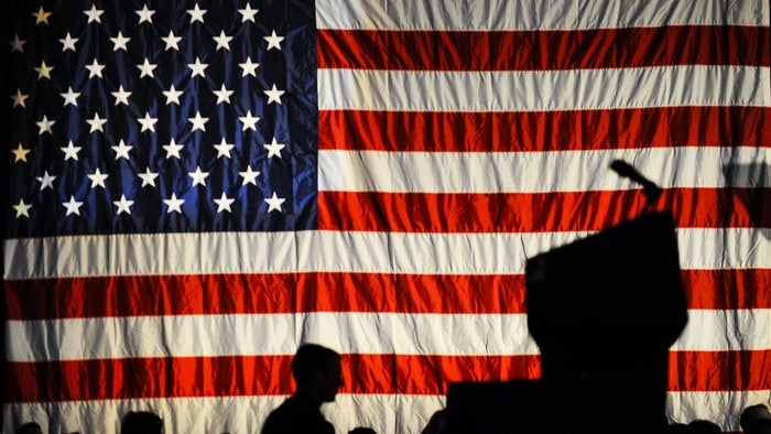 The silhouette of a speaker at a podium is superimposed against an image of an American flag on a stage.