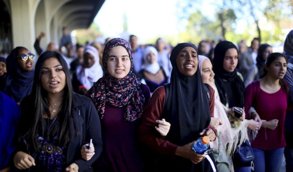 A group of young women stand together with linked arms. Two of the young women wear hijab.