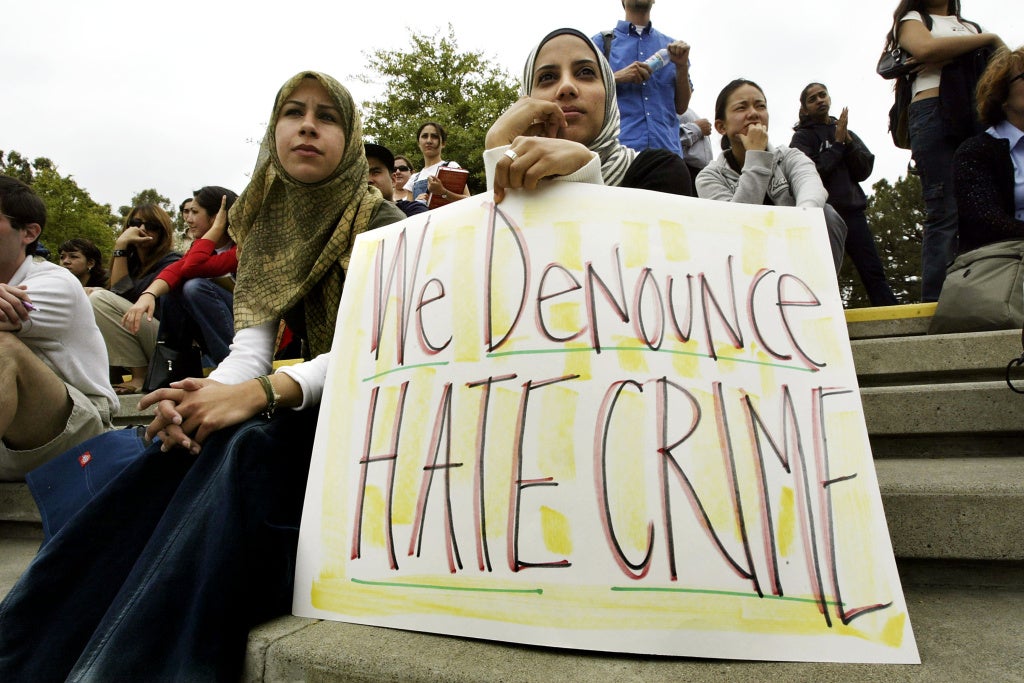 Two people sit on cement steps. The person on the right is holding a paper sign that reads "WE DENOUNCE HATE CRIME" in large handwritten letters.