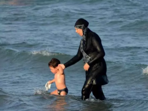 A woman wearing a Burkina walks with a small child in the ocean.