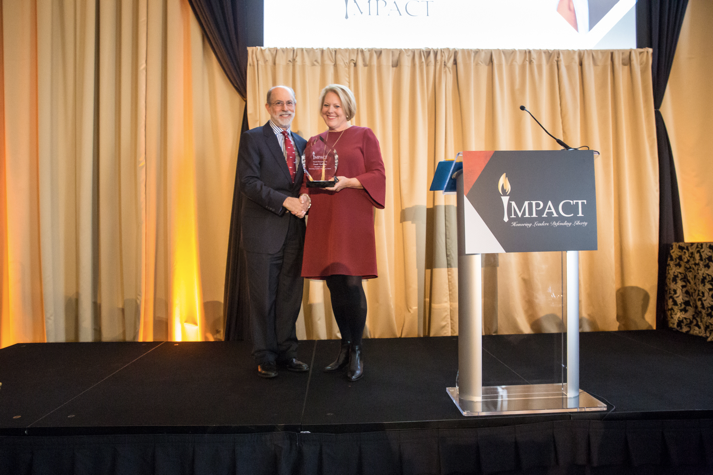 rank Gaffney (left) poses for a picture next to Ginni Thomas (right). Both are standing on a stage, shaking hands and holding an award.