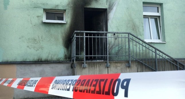 A building that appears to be burned is in the backdrop of a strip of police investigation tape. The tape is labeled in German.