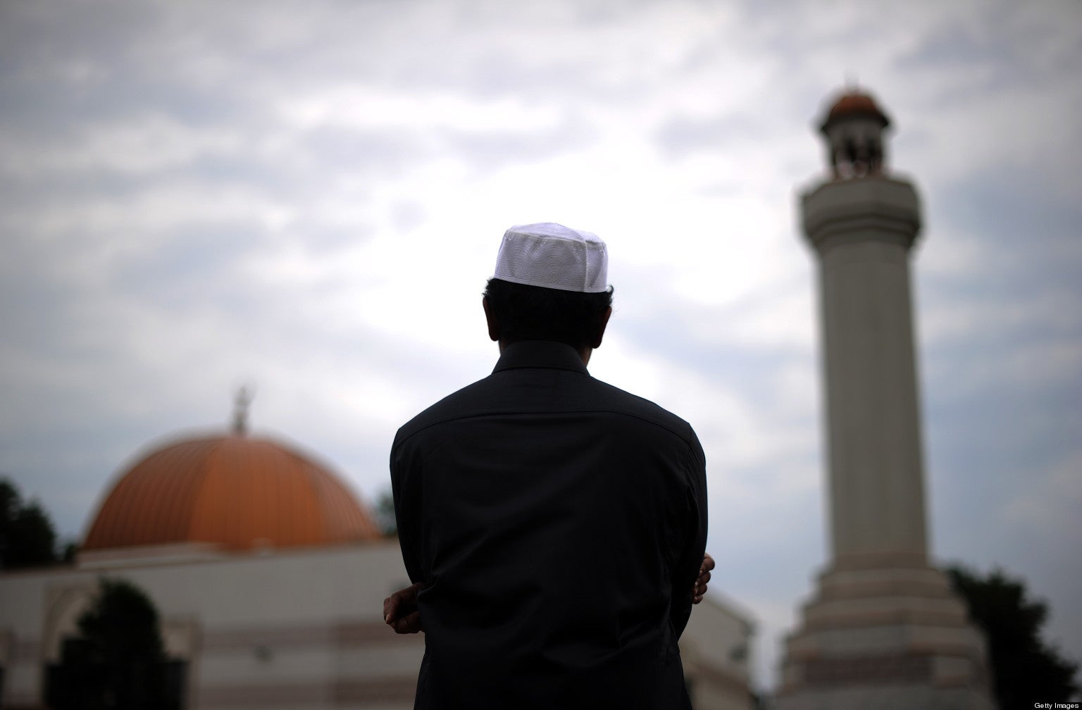 A man looks off at the distance toward a mosque, the image of which is blurred. The man wears a those and a taqiyah.