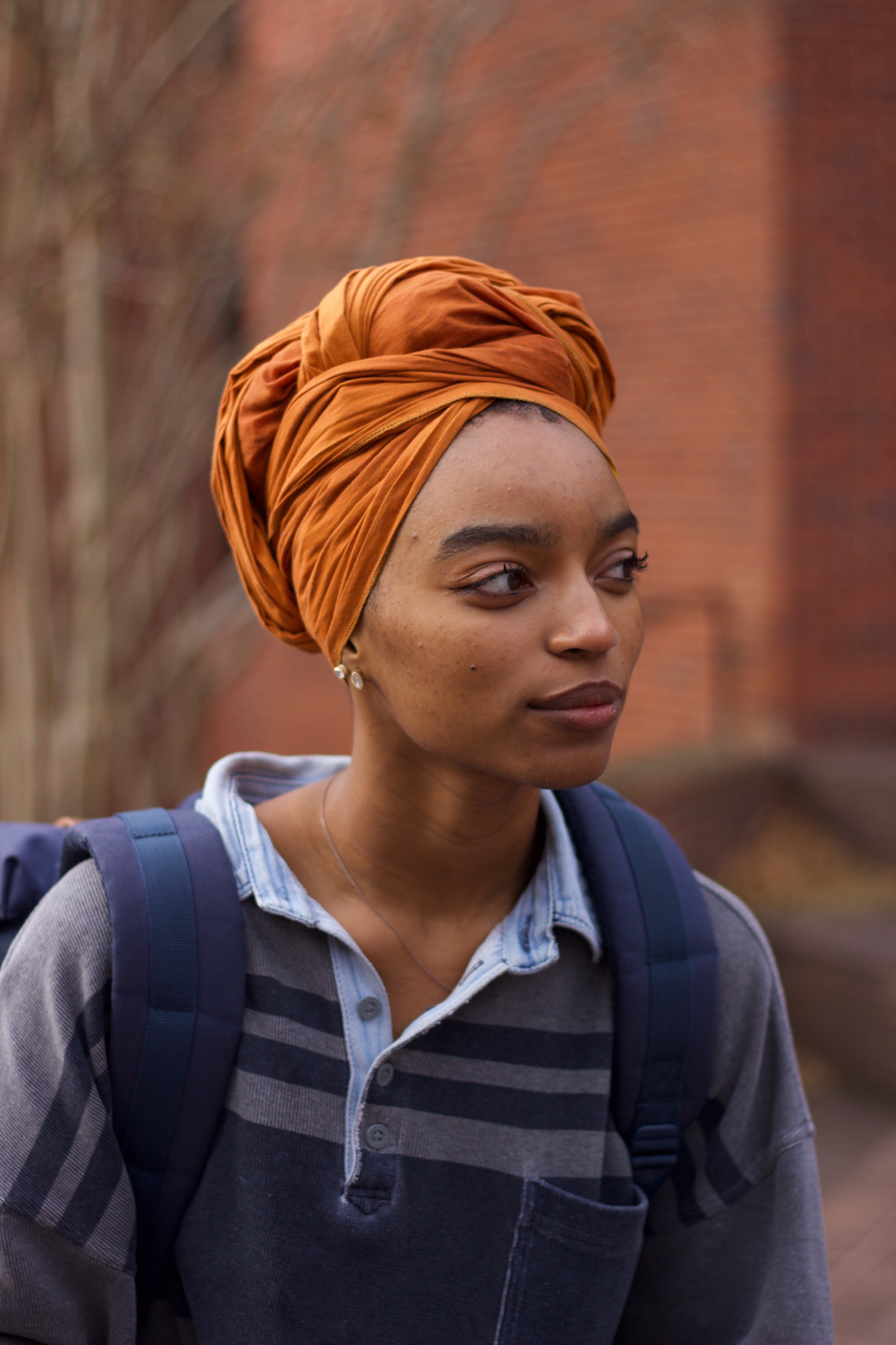 person wearing an orange-colored head scarf, grey and blue-colored polo shirt and blue-colored backpack looks off to the side. The background is a tree without leaves and a brick building