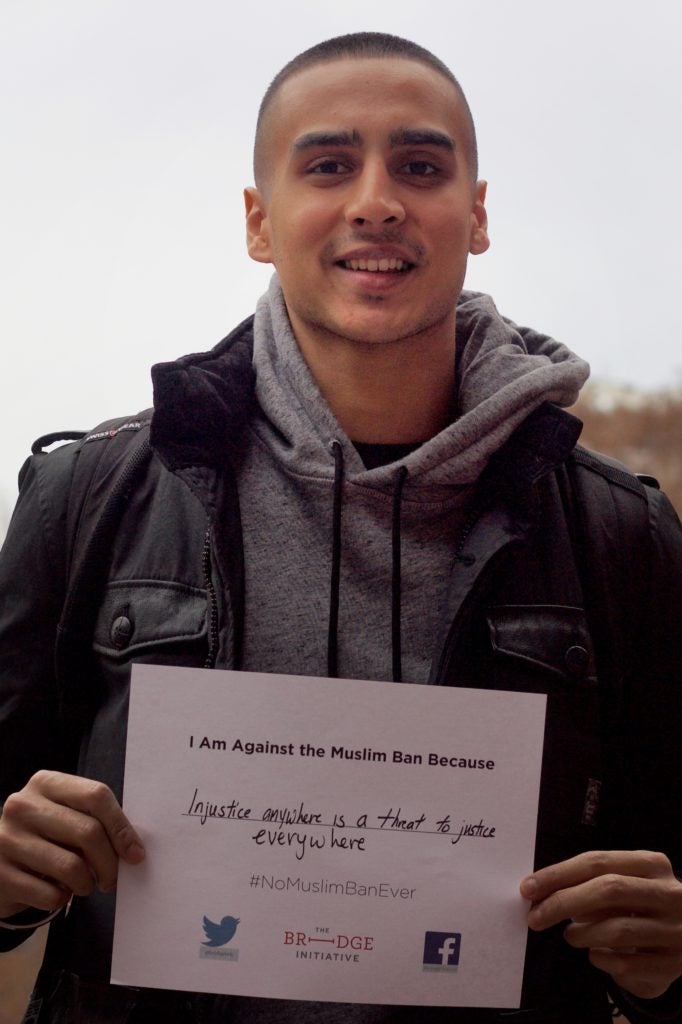 person with short cropped, dark brown hair looks directly in the camera, smiling. The person, who is wearing a grey-colored sweatshirt and black jacket, hold a sign that reads, “I am against the Muslim Ban because injustice anywhere is a threat to justice everywhere.” In the background is an overcast sky and a red-brick building.