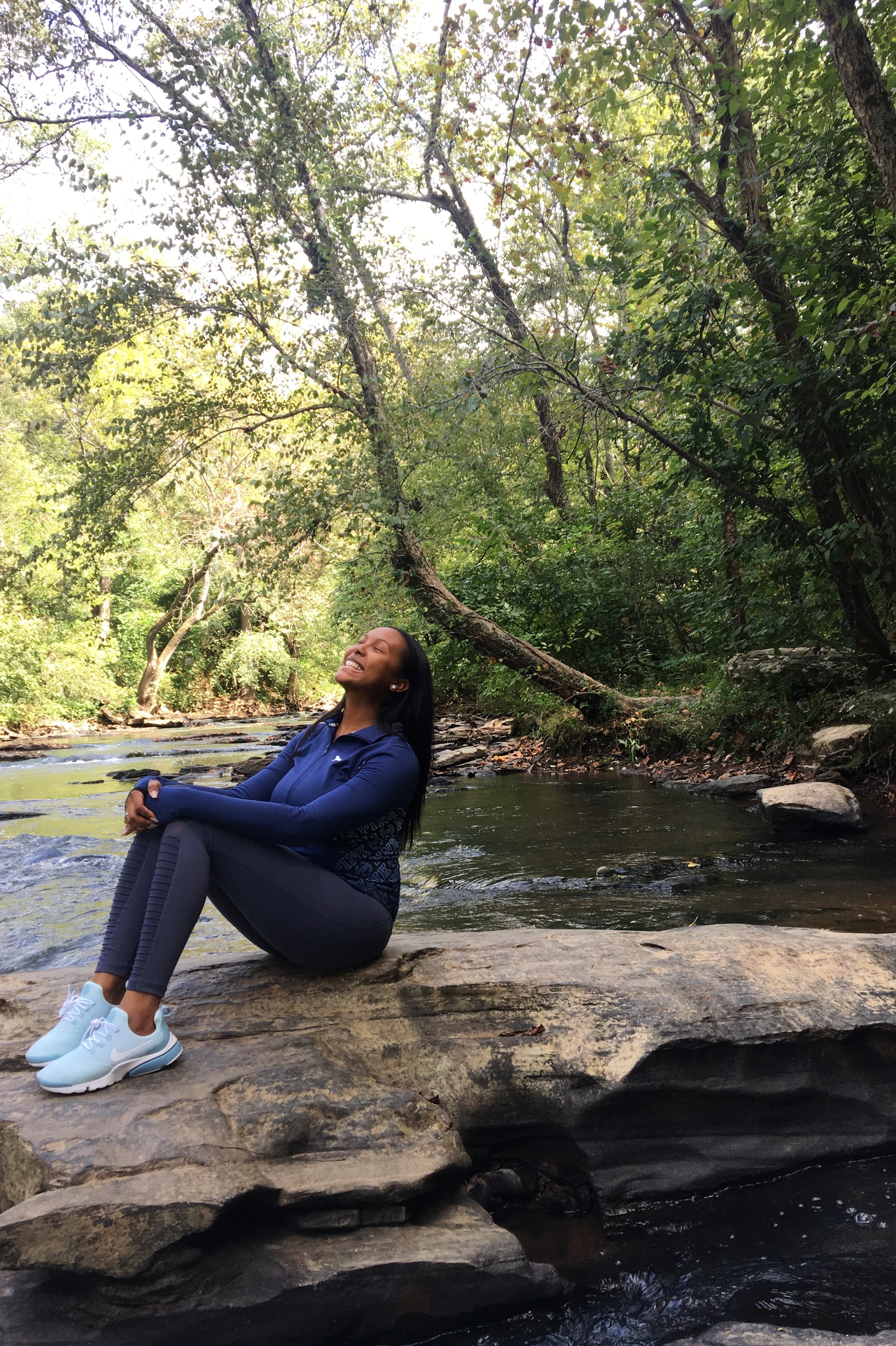 side profile of a person smiling and slightly leaning back while seated on a large rock in the middle of a creek surrounded by trees with green leaves. The person is wearing light-colored sneakers, grey leggings and dark blue long-sleeved shirt.