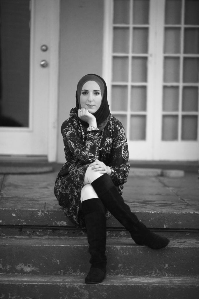 black-and-white photo of a person seated on steps leading up to a house. The person wears knee-high, black-colored boots, light-colored pants, a floral-patterned dress/tunic, a headscarf and a necklace, bracelet and ring. The person looks at the camera, with legs crossed and chin resting in hand.
