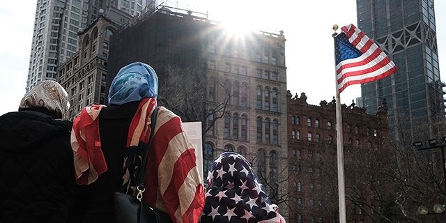 Three women wearing hijab stand with their backs turned to the camera with a high rise building and American flag in the foreground.