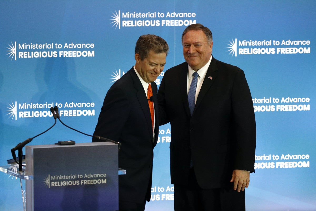 Sam Brownback and Mike Pompeo smile while embracing in a side-hug. In front of them is a podium and behind them is a blue banner with logos that read, "Ministerial to Advance Religious Freedom."