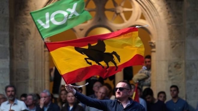 A man in sunglasses waves two flags: a larger flag of Spain and a smaller green flag with the words VOX